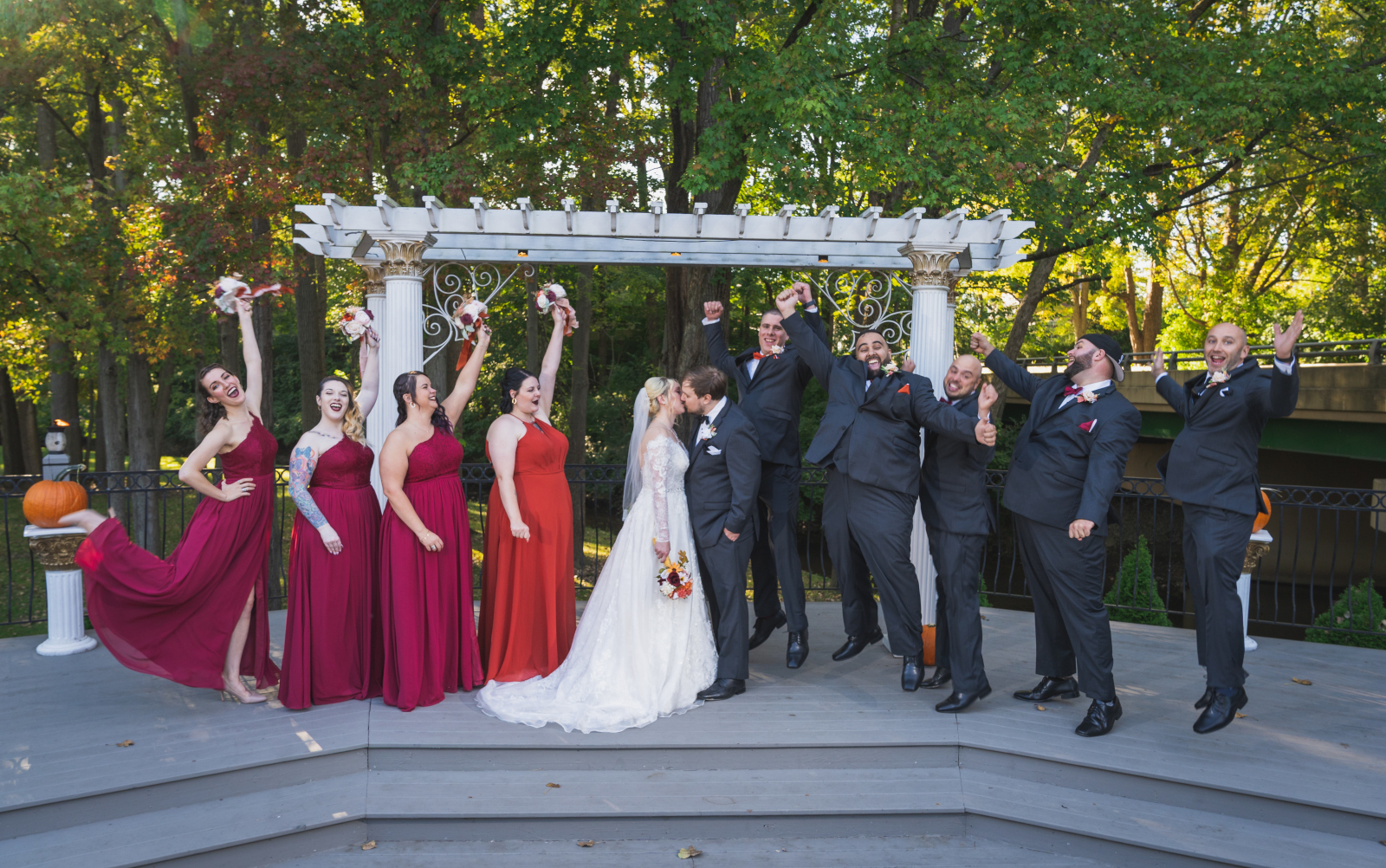 Bride and groom kiss, bridal party cheering, bridal party portrait, cute bridal party portrait, fun bridal party portrait, fun pose, wooden arch with pillars, green, nature, trees, fall wedding, cute outdoor wedding ceremony at Grand Pacific Wedding Gardens