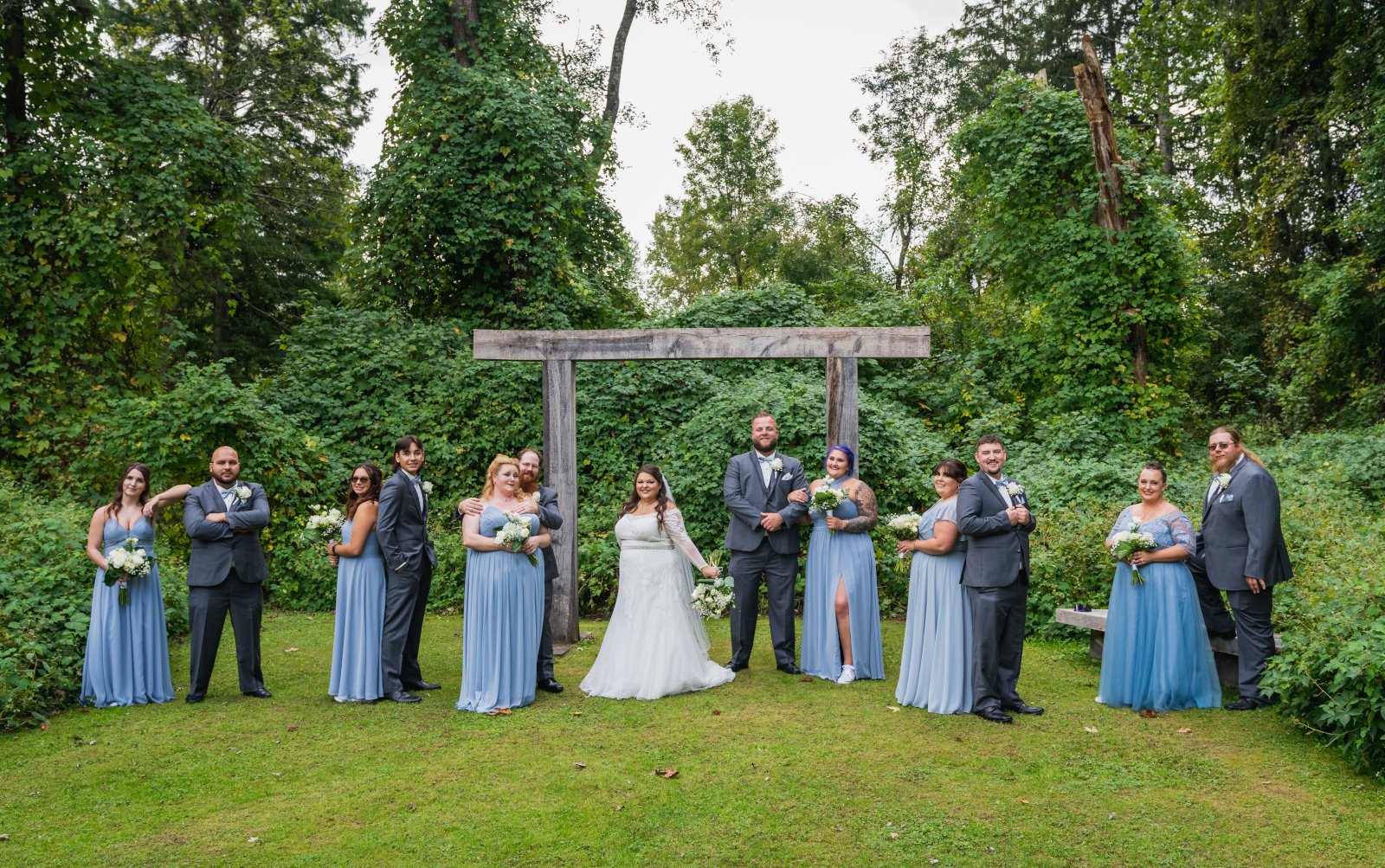 Bride with bridesmaids and groomsmen, bridal party portrait, rustic wooden arch, green, nature, trees, poses for large bridal party, outdoor September wedding ceremony at Westfall Event Center