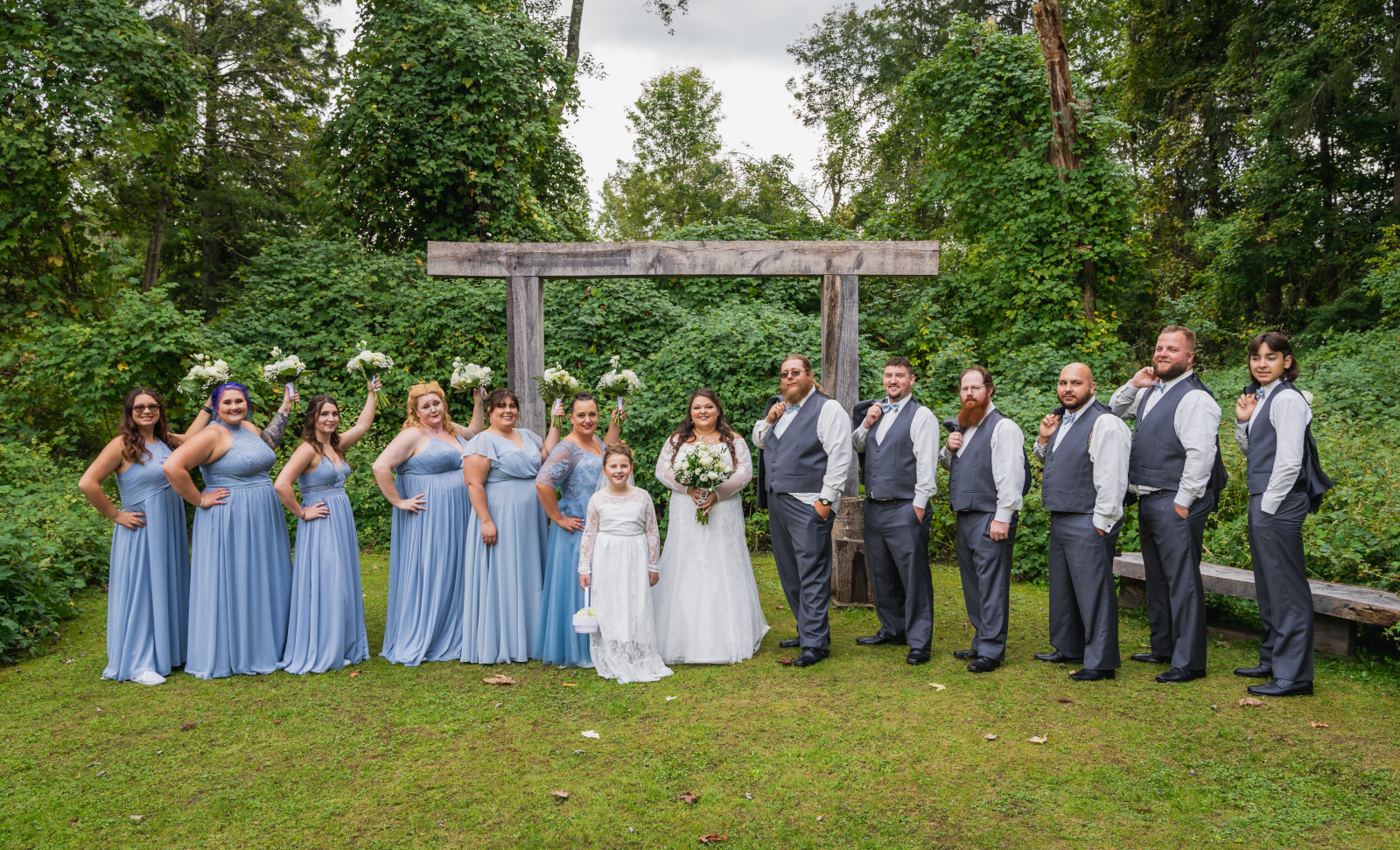 Bride with bridal party, bridesmaids, groomsmen, flower girl, bridal party portrait, flower bouquets in the air, rustic wooden arch, unique bridal party portrait, green, nature, trees, outdoor September wedding ceremony at Westfall Event Center