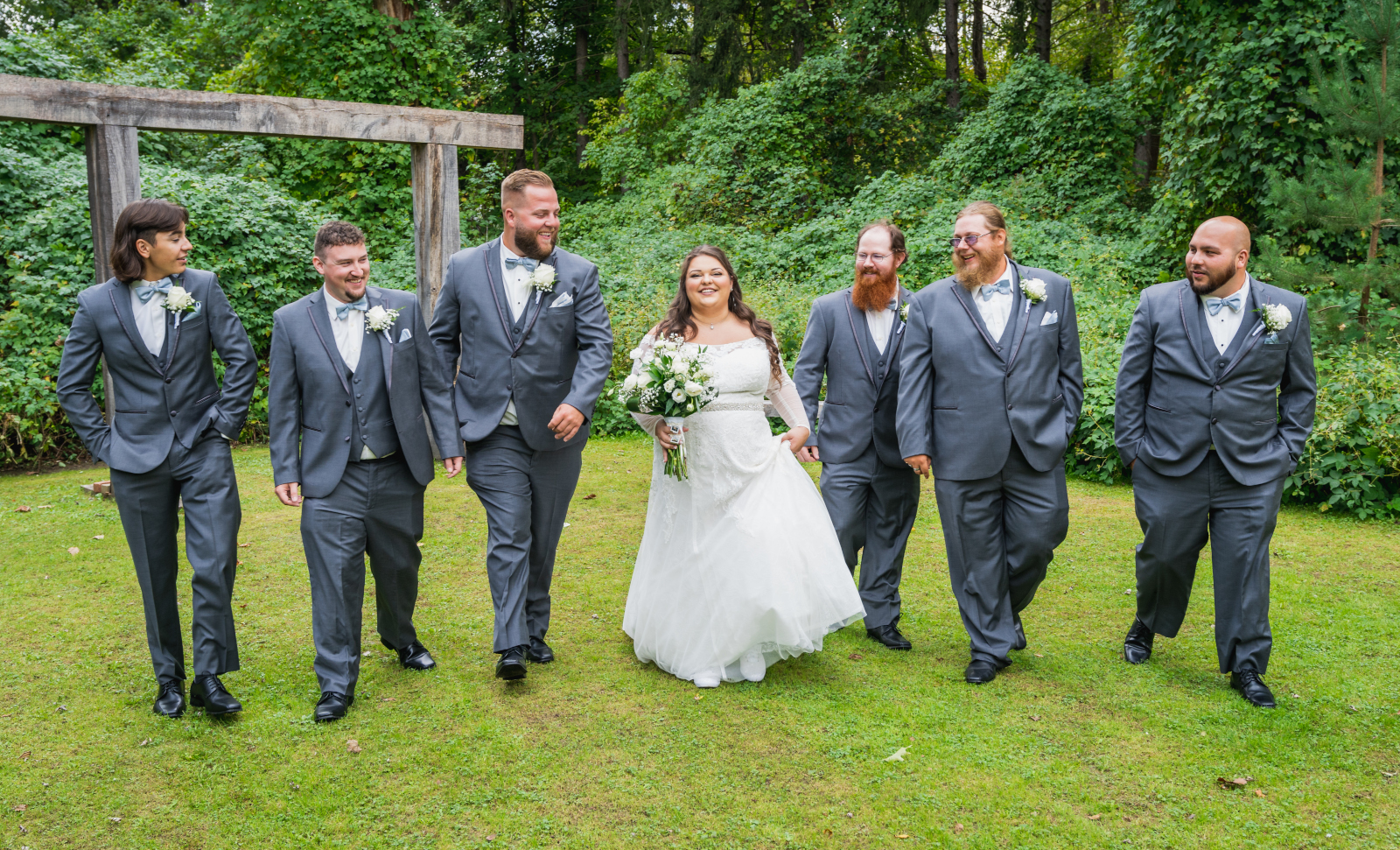 Bride with groomsmen, walking, candid, smiling, rustic wooden arch, green, nature, trees, outdoor September wedding ceremony at Westfall Event Center